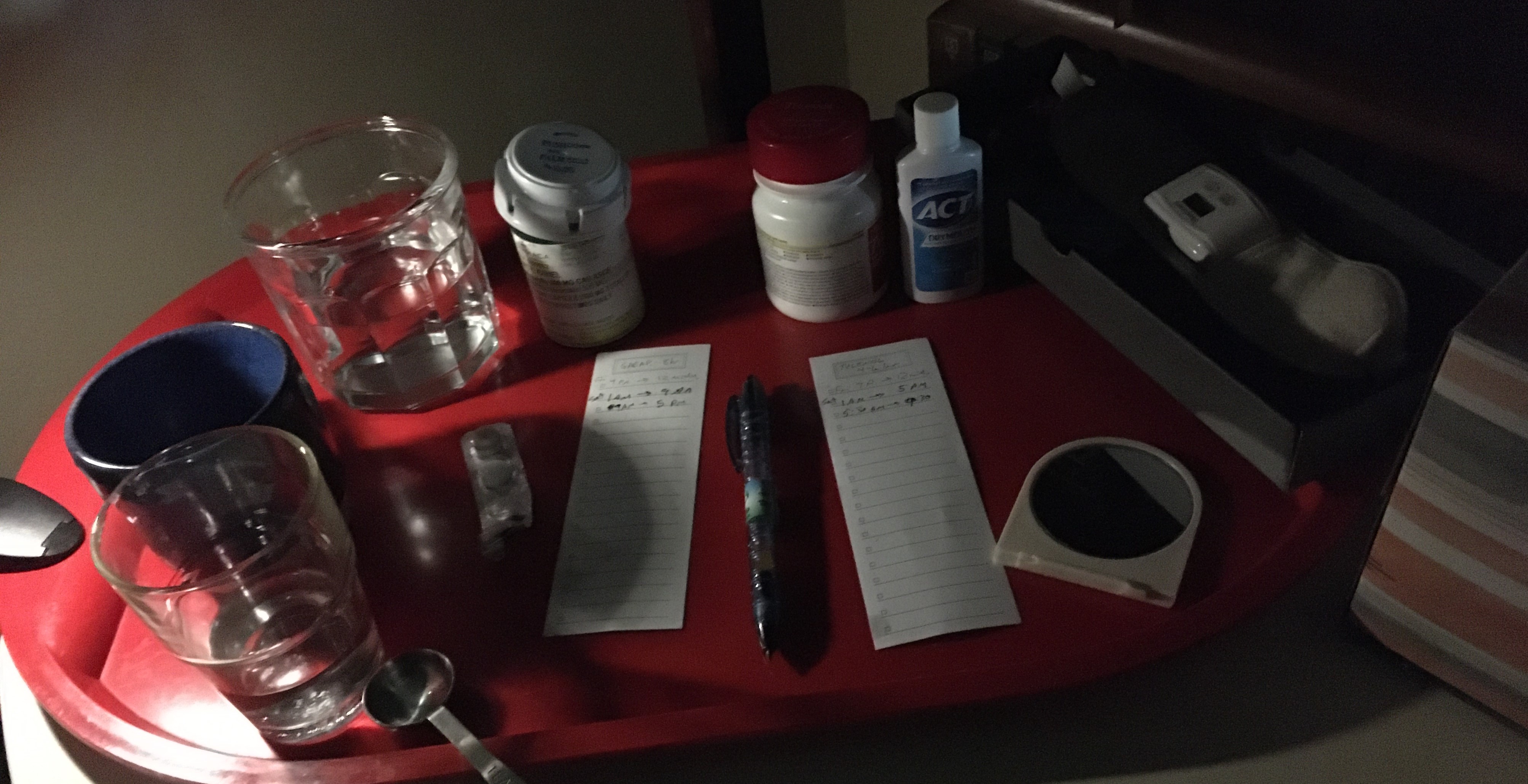 Photo of red tray holding several drinking glasses, bottles of medicine, notes, a mirror.