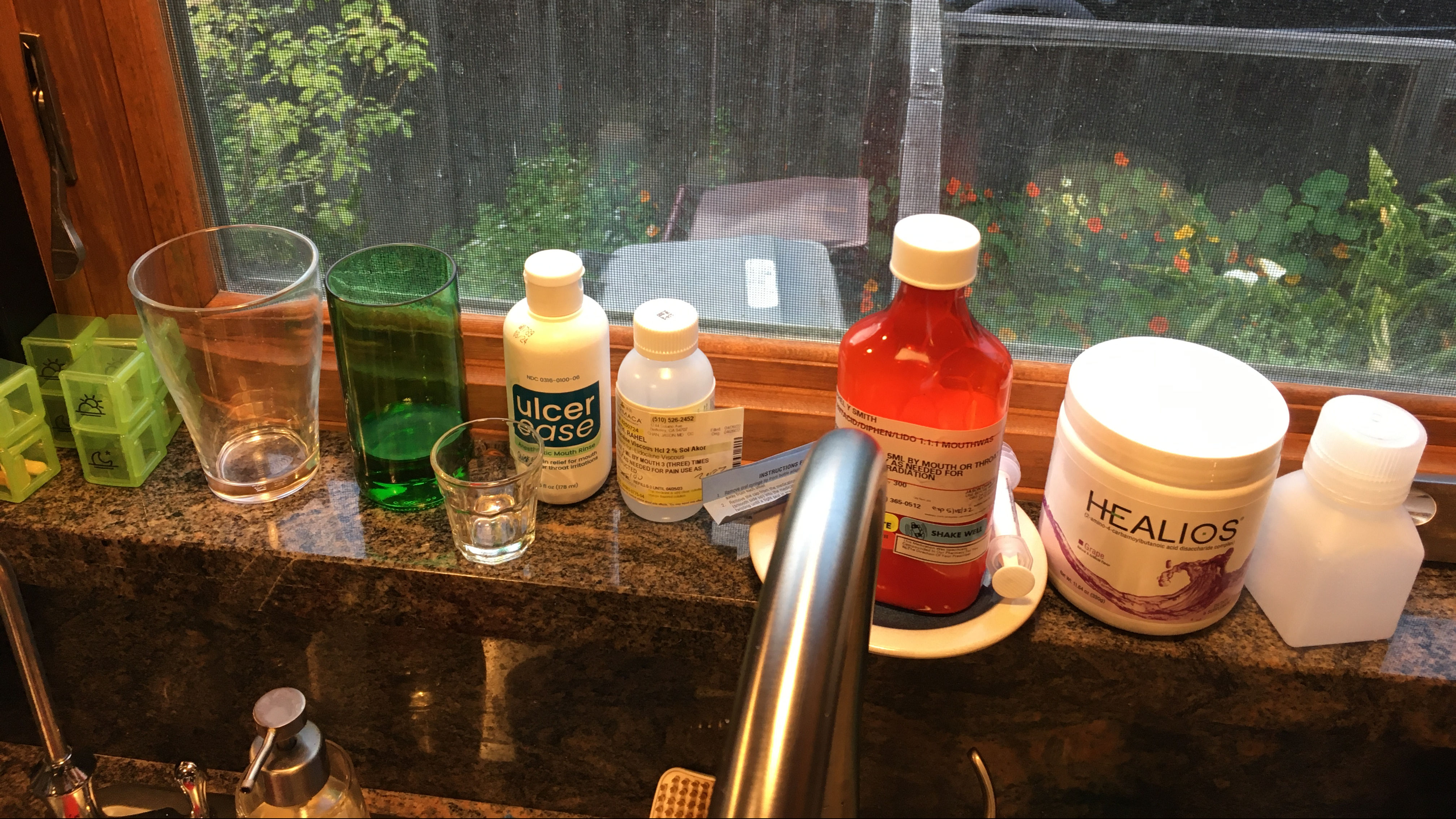 Photo of several pill containers and bottles of numbing solution lined up on a kitchen windowsill, with plant and a fence showing through the window.
