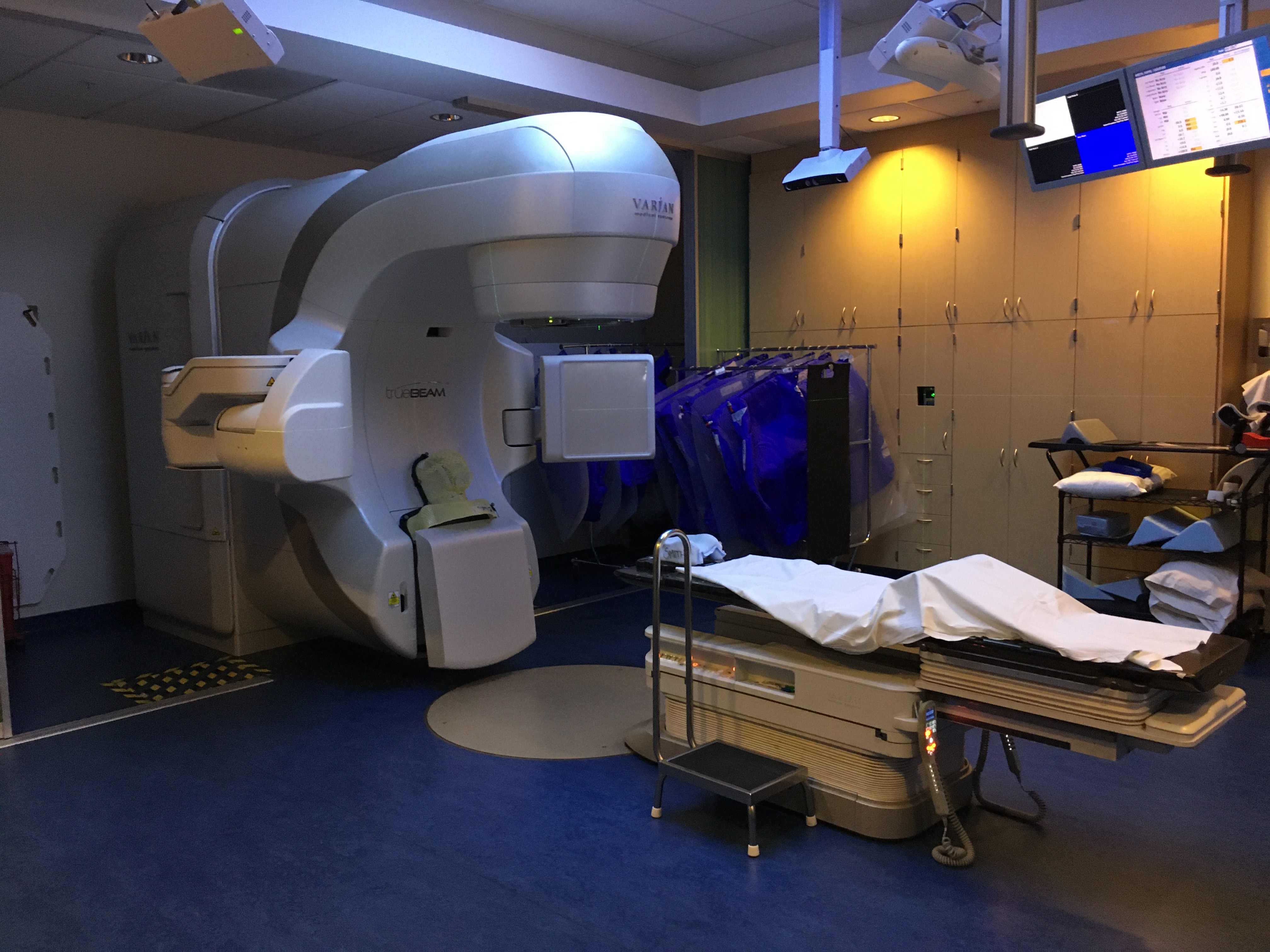 A photo of the radiation treatment room, with the table in the center and the machine.