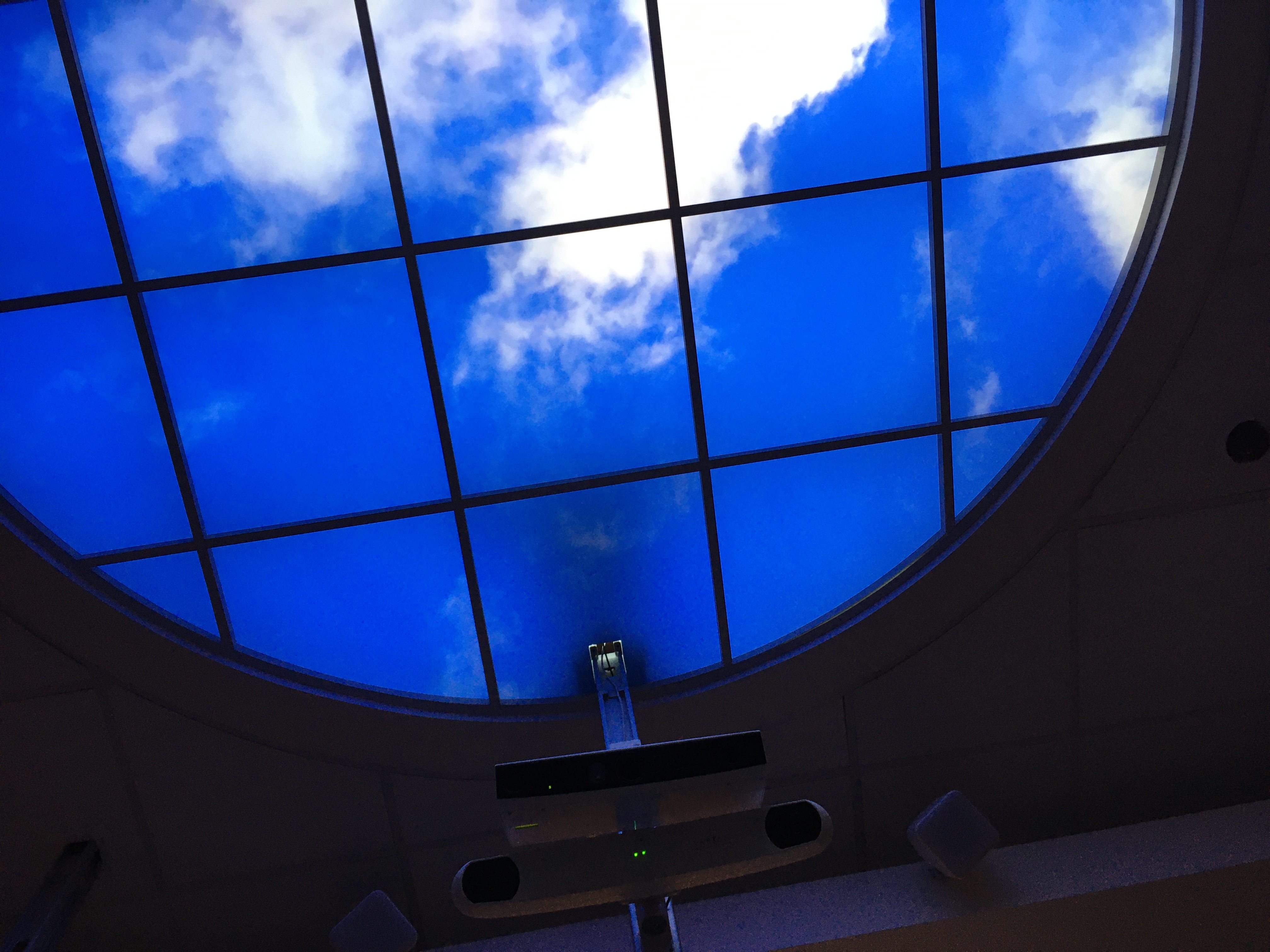 Photo of a ceiling light that looks like a window, with clouds in a blue sky.