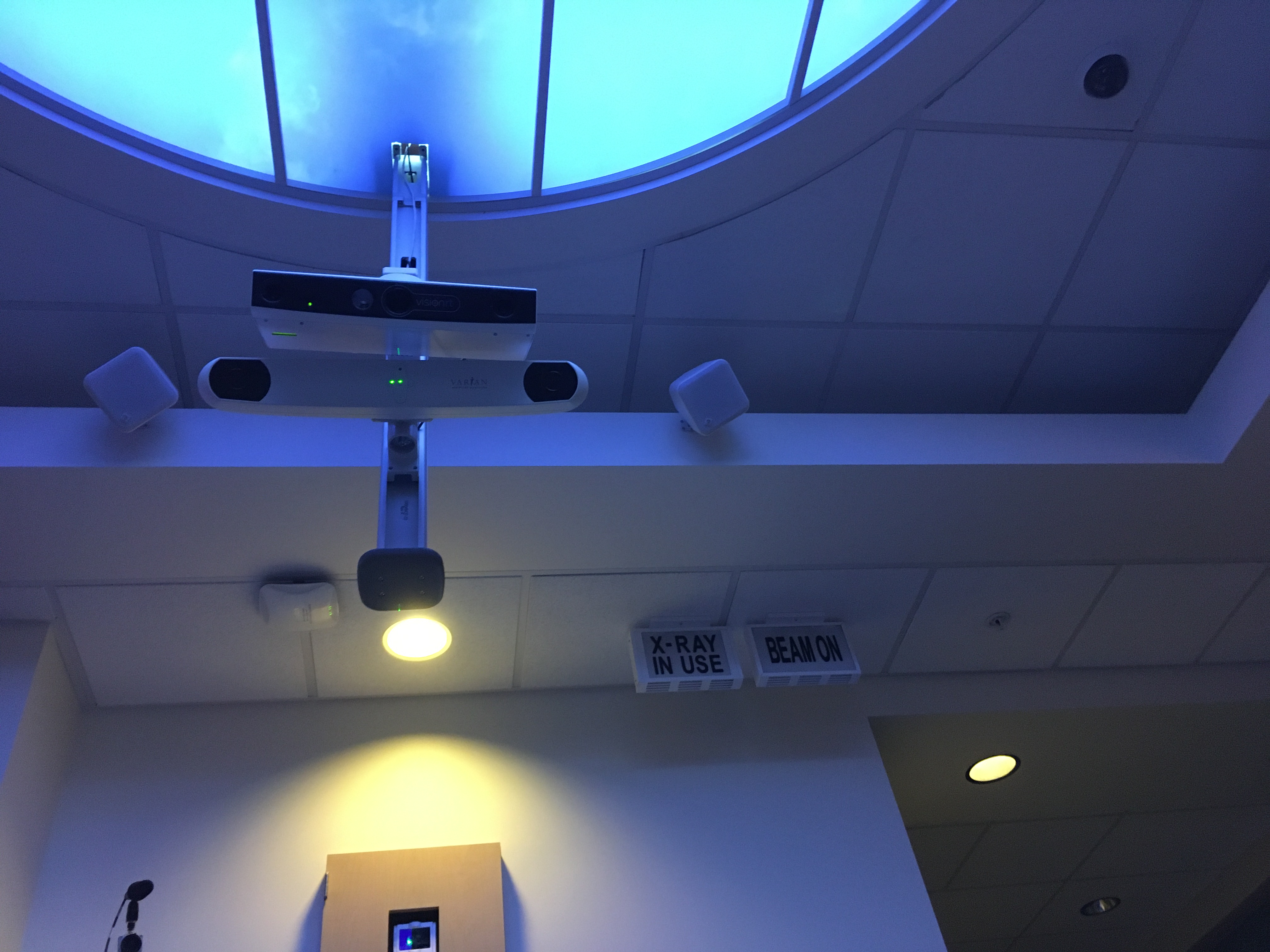Photo of the ceiling and upper wall of a room, with a sky-colored light embedded in the ceiling, several cameras, lights, and/or lasers mounted to the ceiling, and two signs reading "X-Ray in Use" and "Beam On".