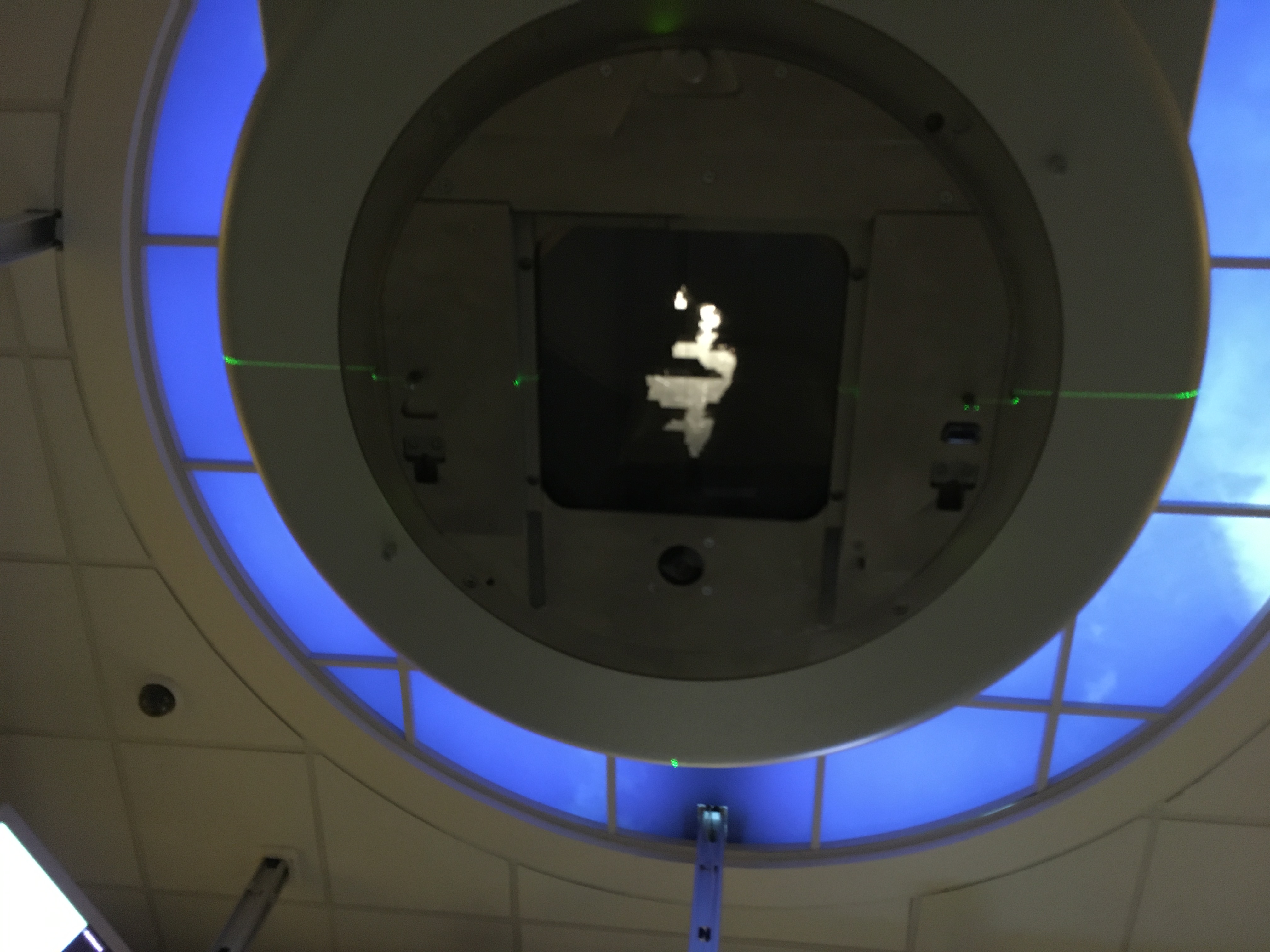 A picture looking up at a radiation machine device, as though from in the machine. There's a shape of light with odd edges shining through a dark rectangle, within circular panel.